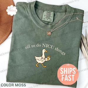 Puede incluir: Una camiseta de cuello redondo color verde musgo con un gr&aacute;fico de un ganso blanco que lleva un estetoscopio y sostiene una taza de caf&eacute;. El texto sobre el ganso dice "off to do NICU things" en letras blancas. Etiqueta Comfort Colors.
