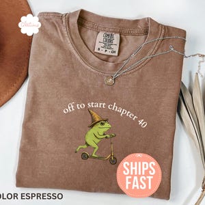 May include: Espresso-colored t-shirt with the text "off to start chapter 40" and a graphic of a green frog riding a scooter. The shirt has a crew neck and short sleeves. A silver necklace and dried plant are also visible.