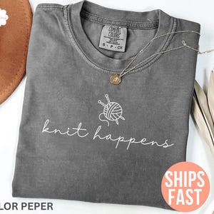 May include: A gray Comfort Colors t-shirt with the words "knit happens" in white script, along with a graphic of yarn and knitting needles. A silver necklace with a pendant rests on the shirt. A brown hat is partially visible.