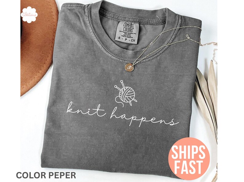 May include: A gray Comfort Colors t-shirt with the words "knit happens" in white script, along with a graphic of yarn and knitting needles. A silver necklace with a pendant rests on the shirt. A brown hat is partially visible.