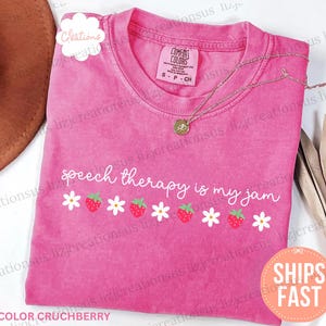 Puede incluir: Una camiseta rosa de manga corta con cuello redondo. La prenda presenta el texto blanco "speech therapy is my jam" sobre una fila horizontal de margaritas blancas y fresas rojas alternadas. La etiqueta interior indica Comfort Colors talla S.