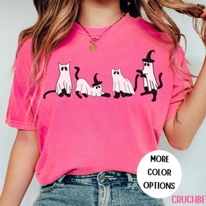 May include: A bright pink t-shirt featuring a playful design of four cartoon cats dressed as ghosts. One cat wears a witch hat. The shirt has the text "MORE COLOR OPTIONS" and the brand name "CRUCHBERRY" in a white circle.