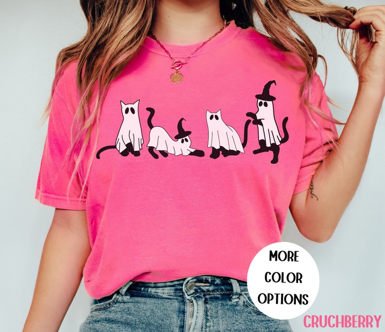 May include: A bright pink t-shirt featuring a playful design of four cartoon cats dressed as ghosts. One cat wears a witch hat. The shirt has the text "MORE COLOR OPTIONS" and the brand name "CRUCHBERRY" in a white circle.