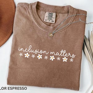 May include: Espresso-colored t-shirt with the phrase "inclusion matters" in white script, decorated with small daisy graphics. A silver necklace with a pendant rests on the shirt. The shirt is folded, with a brown hat visible in the background.