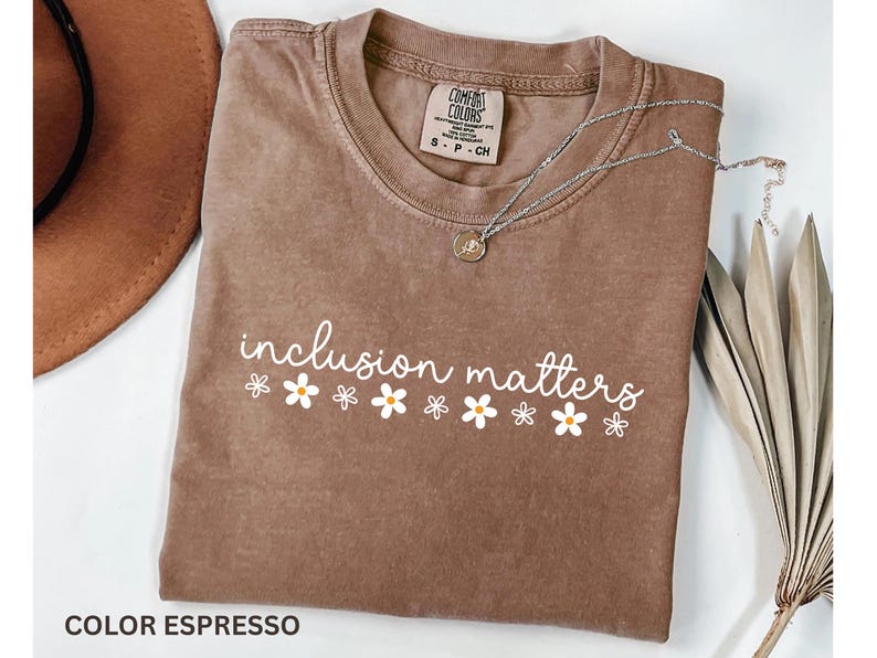 May include: Espresso-colored t-shirt with the phrase "inclusion matters" in white script, decorated with small daisy graphics. A silver necklace with a pendant rests on the shirt. The shirt is folded, with a brown hat visible in the background.