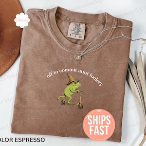 May include: Espresso-colored t-shirt with a whimsical frog graphic riding a scooter, wearing a wizard hat. The text "off to commit aunt foolery" is above the graphic. A silver necklace and a brown hat are also visible.