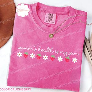 May include: A pink crew neck t-shirt with a vintage wash finish. The front features the white text "women's health is my jam" above a horizontal row of alternating white daisies and red strawberries. A gold-toned necklace rests on the collar.