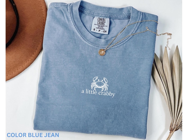 May include: A blue jean colored t-shirt with the words "a little crabby" and a crab graphic. The shirt has a small tag on the collar and is paired with a gold necklace. A brown hat and dried plant are in the background.