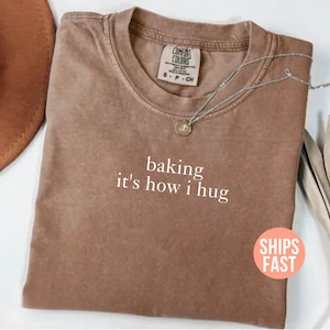 Może przedstawiać: Brązowa koszulka z białym napisem "baking it's how I hug". Koszulka ma okrągły dekolt i krótkie rękawy. Srebrny naszyjnik z wisiorkiem jest ułożony na koszulce.
