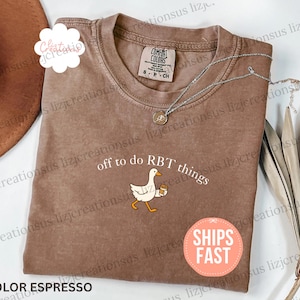 May include: An espresso-colored short-sleeve t-shirt with a crew neckline. The shirt features a graphic of a white goose holding a coffee cup, accompanied by the white text "off to do RBT things" printed in a simple, clean font above the goose.