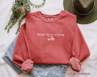 May include: A crimson long-sleeve crewneck sweatshirt featuring the white text "wee woo crew" centered above a small white line-art graphic of an ambulance. The sweatshirt has a ribbed collar, cuffs, and hem, with a soft, fleece-like interior texture.
