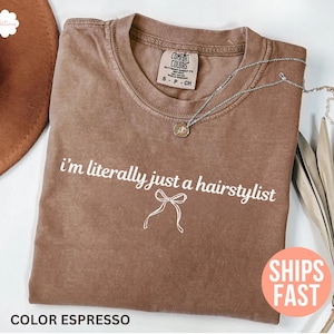 May include: A brown t-shirt with the phrase "I'm literally just a hairstylist" printed in white. A silver necklace with a pendant is on the shirt. The t-shirt has a crew neck and a relaxed fit.