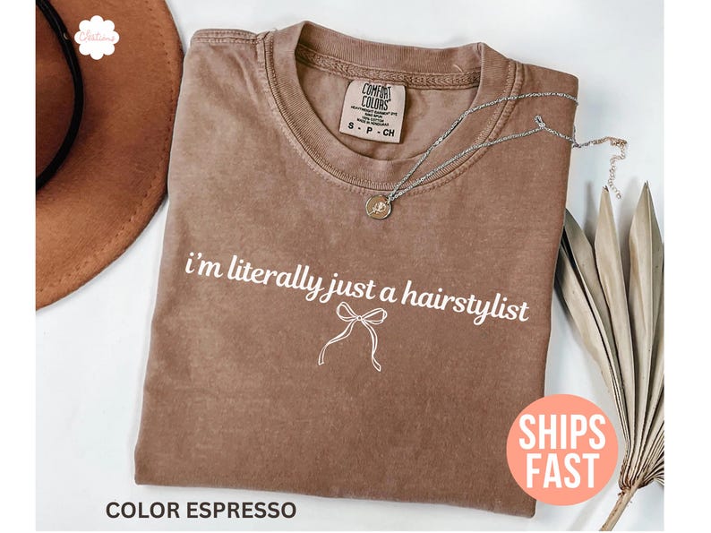 May include: A brown t-shirt with the phrase "I'm literally just a hairstylist" printed in white. A silver necklace with a pendant is on the shirt. The t-shirt has a crew neck and a relaxed fit.