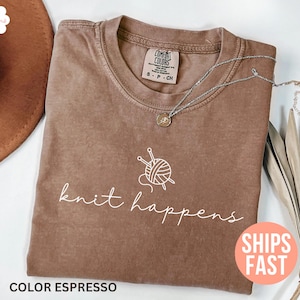 May include: Espresso-colored t-shirt with the phrase "knit happens" in white script. A white graphic of yarn and knitting needles is above the text. A silver necklace with a small pendant is on the shirt.