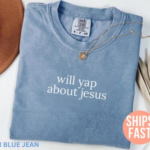 May include: A blue jean colored t-shirt with the phrase "will yap about jesus" in white lettering. A gold necklace with a circular pendant rests on the shirt. The shirt is folded, with a brown hat and dried plant in the background.