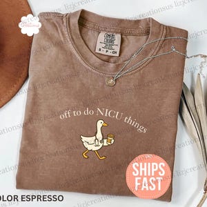 Puede incluir: Una camiseta marr&oacute;n de manga corta con el gr&aacute;fico de un ganso blanco que lleva un estetoscopio y sostiene una taza de caf&eacute;. El texto sobre el ganso dice off to do NICU things. La camiseta tiene una etiqueta Comfort Colors en el cuello.