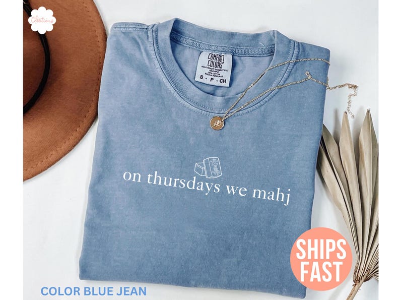 以下が含まれることがあります： 「on thursdays we mahj」の白文字が入った、ブルーデニムカラーのTシャツ。 金色のネックレスがシャツの上に置かれています。 シャツは折りたたまれており、「Comfort Colors」と書かれたタグが付いています。