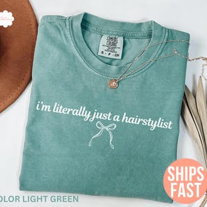 May include: Light green t-shirt with the text "i'm literally just a hairstylist" in white script. A small bow graphic is below the text. A gold necklace with a pendant is on the shirt. The shirt is folded and has a Comfort Colors tag.