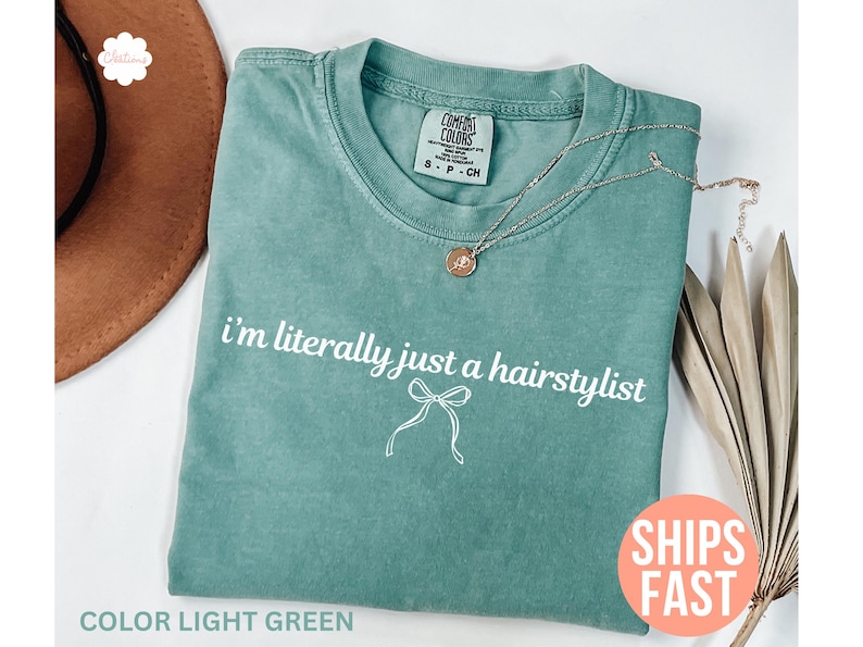 May include: Light green t-shirt with the text "i'm literally just a hairstylist" in white script. A small bow graphic is below the text. A gold necklace with a pendant is on the shirt. The shirt is folded and has a Comfort Colors tag.