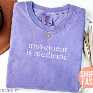 K&ouml;nnte beinhalten: Ein fliederfarbenes Comfort Colors T-Shirt mit dem wei&szlig;en Schriftzug "movement is medicine". Eine goldene Halskette mit Anh&auml;nger liegt auf dem Shirt. Ein brauner Hut und getrocknete Palmbl&auml;tter sind im Hintergrund.