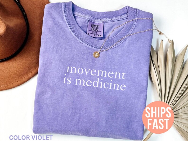 K&ouml;nnte beinhalten: Ein fliederfarbenes Comfort Colors T-Shirt mit dem wei&szlig;en Schriftzug "movement is medicine". Eine goldene Halskette mit Anh&auml;nger liegt auf dem Shirt. Ein brauner Hut und getrocknete Palmbl&auml;tter sind im Hintergrund.