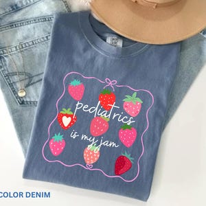 May include: A denim blue t-shirt with a strawberry design and the text "pediatrics is my jam." The shirt is paired with blue jeans and a tan hat. The design features several strawberries in various shades of red and pink.