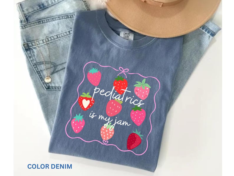 May include: A denim blue t-shirt with a strawberry design and the text "pediatrics is my jam." The shirt is paired with blue jeans and a tan hat. The design features several strawberries in various shades of red and pink.
