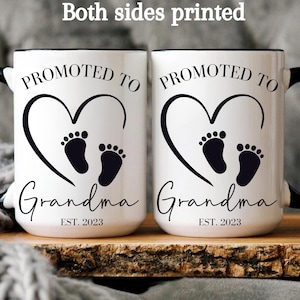 K&ouml;nnte beinhalten: Zwei wei&szlig;e Keramikbecher mit schwarzen Henkeln. Beide Becher zeigen ein schwarzes Herz mit zwei Baby-Fu&szlig;abdr&uuml;cken darin. Der Text "PROMOTED TO GRANDMA EST. 2023" ist auf jedem Becher aufgedruckt.