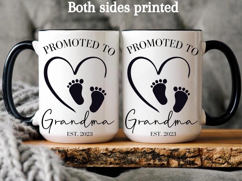 K&ouml;nnte beinhalten: Zwei wei&szlig;e Keramikbecher mit schwarzen Henkeln. Beide Becher zeigen ein schwarzes Herz mit zwei Baby-Fu&szlig;abdr&uuml;cken darin. Der Text "PROMOTED TO GRANDMA EST. 2023" ist auf jedem Becher aufgedruckt.