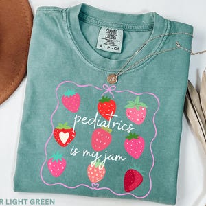 May include: Light green t-shirt with a strawberry design and the text "pediatrics is my jam". The shirt has a crew neck and short sleeves. A gold necklace with a pendant is on top of the shirt.