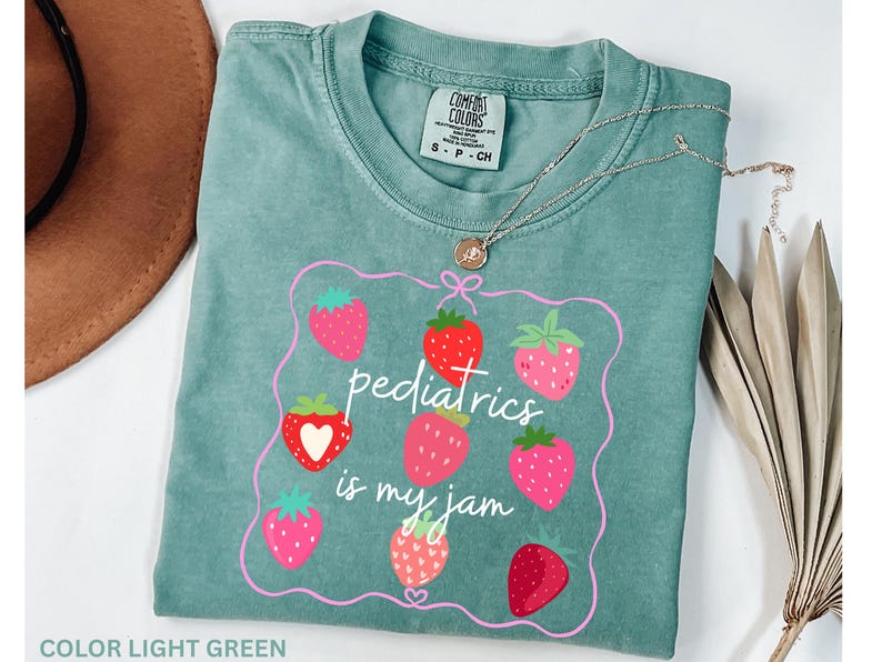 May include: Light green t-shirt with a strawberry design and the text "pediatrics is my jam". The shirt has a crew neck and short sleeves. A gold necklace with a pendant is on top of the shirt.