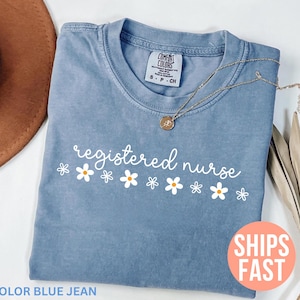 Könnte beinhalten: Blaues Jeans-T-Shirt mit dem Schriftzug "registered nurse" in weißer Schrift, verziert mit kleinen Gänseblümchen-Grafiken. Eine goldene Halskette mit Anhänger liegt auf dem Shirt. Das Shirt ist gefaltet, mit einem braunen Hut im Hintergrund.