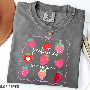 May include: A grey Comfort Colors t-shirt featuring a strawberry design and the text "pediatrics is my jam" in pink. The shirt has a crew neck and short sleeves. A silver necklace with a small pendant is on the shirt.