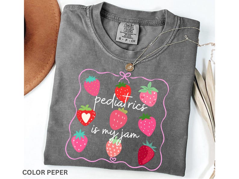 May include: A grey Comfort Colors t-shirt featuring a strawberry design and the text "pediatrics is my jam" in pink. The shirt has a crew neck and short sleeves. A silver necklace with a small pendant is on the shirt.