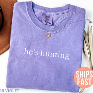 May include: A violet-colored t-shirt with the phrase "he's hunting" printed in white. A gold necklace with a pendant rests on the shirt. A brown hat and dried palm leaves are in the background.