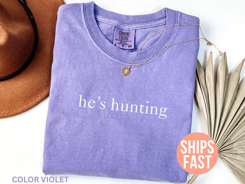 May include: A violet-colored t-shirt with the phrase "he's hunting" printed in white. A gold necklace with a pendant rests on the shirt. A brown hat and dried palm leaves are in the background.