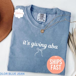 May include: A blue jean colored t-shirt with a crew neckline and short sleeves. The front features the white text it's giving aba above a simple white line drawing of a bow. The shirt has a Comfort Colors brand label at the inner neck.
