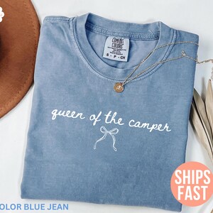May include: Blue jean Comfort Colors t-shirt with the text "queen of the camper" in white script. A gold necklace with a pendant rests on the shirt. A brown hat and dried leaves are in the background.