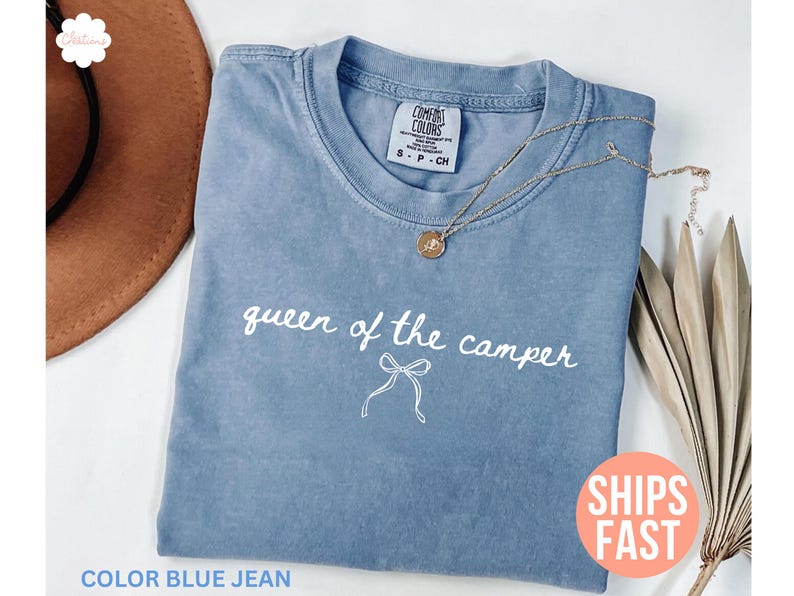 May include: Blue jean Comfort Colors t-shirt with the text "queen of the camper" in white script. A gold necklace with a pendant rests on the shirt. A brown hat and dried leaves are in the background.