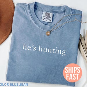 May include: A blue jean colored t-shirt with the phrase "he's hunting" printed in white. A gold necklace with a pendant rests on the shirt. A brown hat and dried plant are in the background.