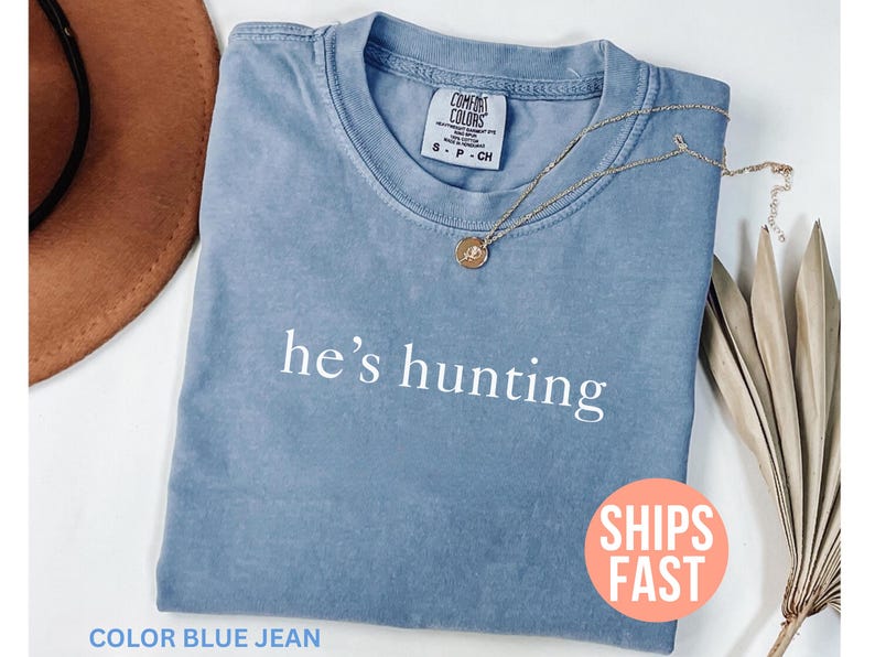 May include: A blue jean colored t-shirt with the phrase "he's hunting" printed in white. A gold necklace with a pendant rests on the shirt. A brown hat and dried plant are in the background.