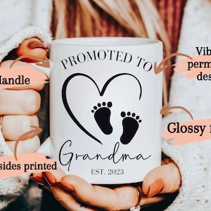 K&ouml;nnte beinhalten: Wei&szlig;e Keramik-Kaffeetasse mit einem schwarzen Design, das "ZUR OMA BEF&Ouml;Rdert EST. 2023" mit einem Herzen und zwei Baby-Fu&szlig;abdr&uuml;cken im Herzen sagt. Die Tasse hat einen C-f&ouml;rmigen Griff und eine gl&auml;nzende Oberfl&auml;che.