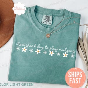 May include: Light green t-shirt with the phrase "it's a great day to play mahjong" in white script, decorated with daisy graphics. A gold necklace rests on the shirt. The shirt is a Comfort Colors brand.