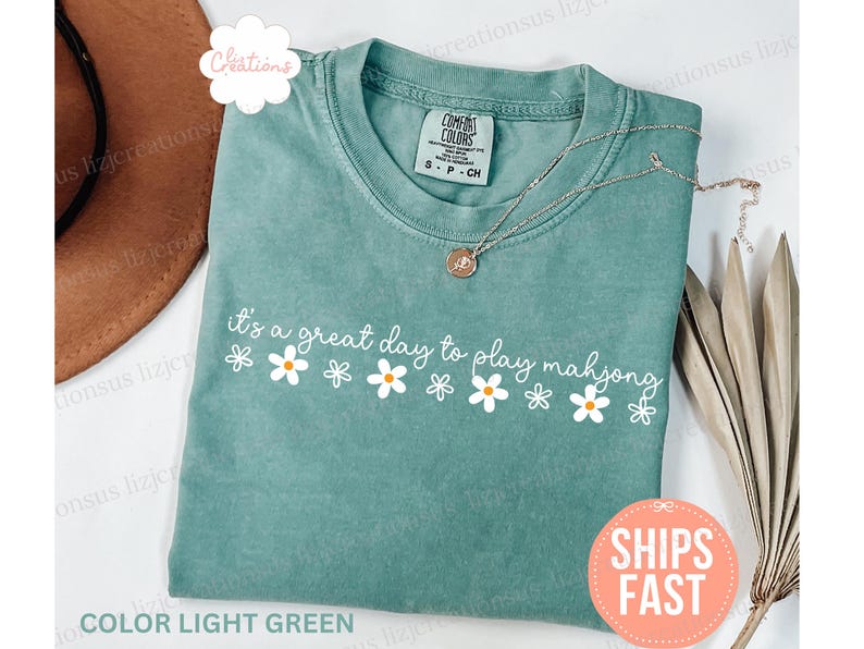 May include: Light green t-shirt with the phrase "it's a great day to play mahjong" in white script, decorated with daisy graphics. A gold necklace rests on the shirt. The shirt is a Comfort Colors brand.