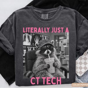 May include: A charcoal grey t-shirt featuring a graphic of a raccoon wearing a baseball cap and sunglasses while holding an iced coffee. The text LITERALLY JUST A CT TECH is printed in pink above and below the black and white image.