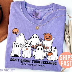 May include: A lavender Comfort Colors t-shirt featuring a graphic of cartoon ghosts with pumpkins and flowers. The text "DON'T GHOST YOUR FEELINGS talk about them" is printed below the ghosts. A gold necklace is draped on the shirt.