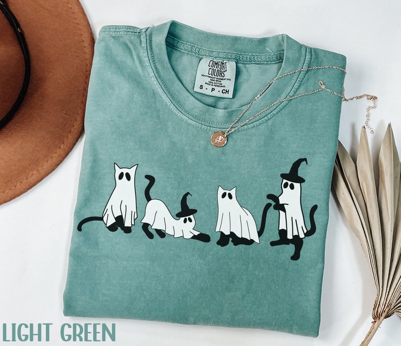 May include: Light green t-shirt with a graphic of four cartoon cats dressed as ghosts, one wearing a witch hat. The shirt has a crew neck and short sleeves. A silver necklace with a pendant is draped across the shirt. The text "LIGHT GREEN" is at the bottom.