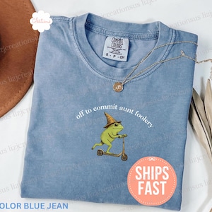 May include: A blue jean colored t-shirt with the phrase "off to commit aunt foolery" and a graphic of a frog on a scooter. The shirt has a round neck and short sleeves. A gold necklace is draped across the shirt.