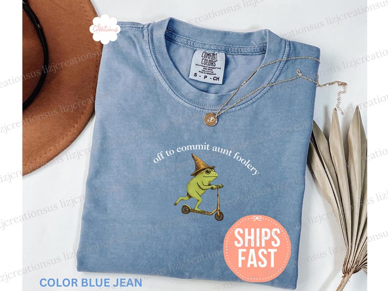 May include: A blue jean colored t-shirt with the phrase "off to commit aunt foolery" and a graphic of a frog on a scooter. The shirt has a round neck and short sleeves. A gold necklace is draped across the shirt.