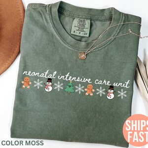 May include: Olive green t-shirt with the text "neonatal intensive care unit" and festive gingerbread men, snowmen, and Christmas trees. The shirt has a crew neck and short sleeves. The text "COLOR MOSS" is at the bottom.